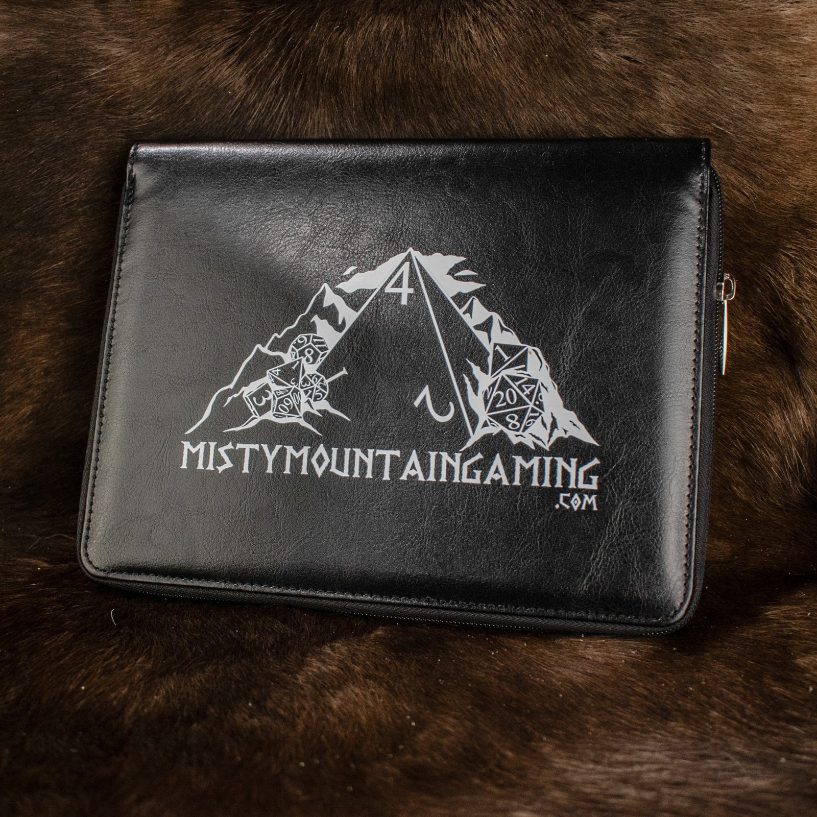 A black poly-leather zippered dice case with foam lining, featuring a white logo 'Mistymountaingaming.com' on the front, placed on a furry surface.