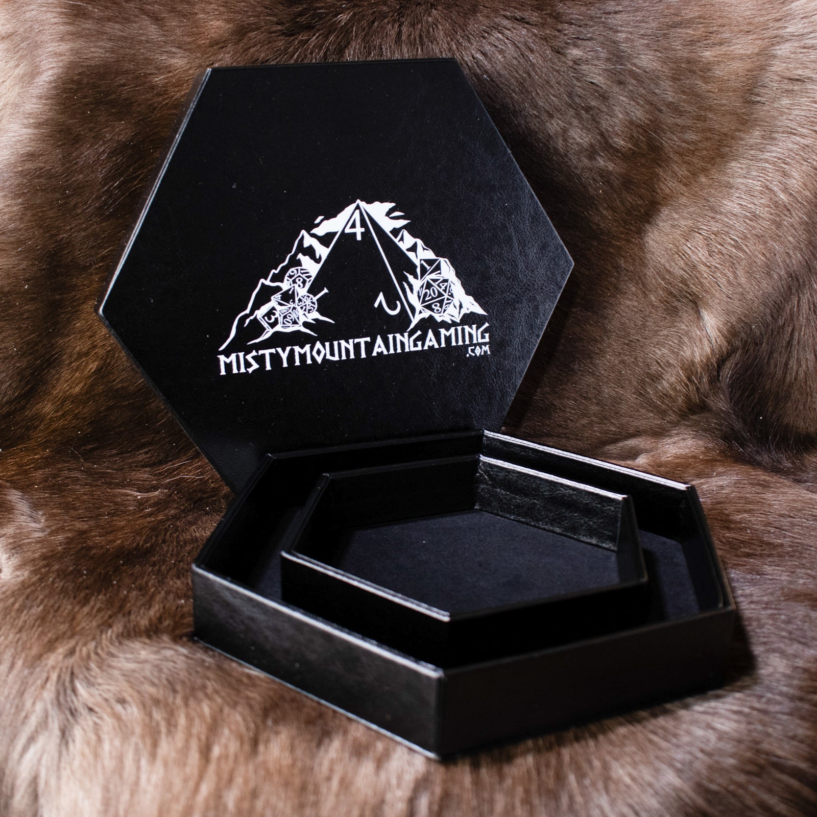 A black hexagonal dice tray with a misty mountain gaming logo, placed on a furry surface.