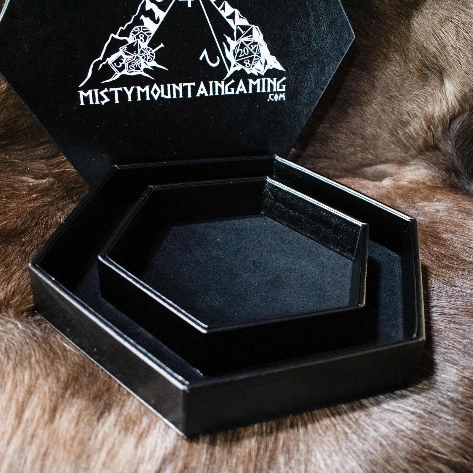 A black hexagonal dice tray with a misty mountain gaming logo, placed on a furry surface.