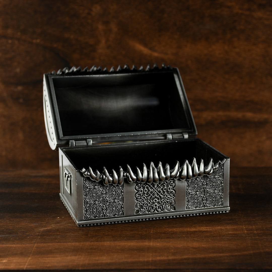 A silver metal chest designed to look like a mimic with teeth, open and filled with multicolored gaming dice.