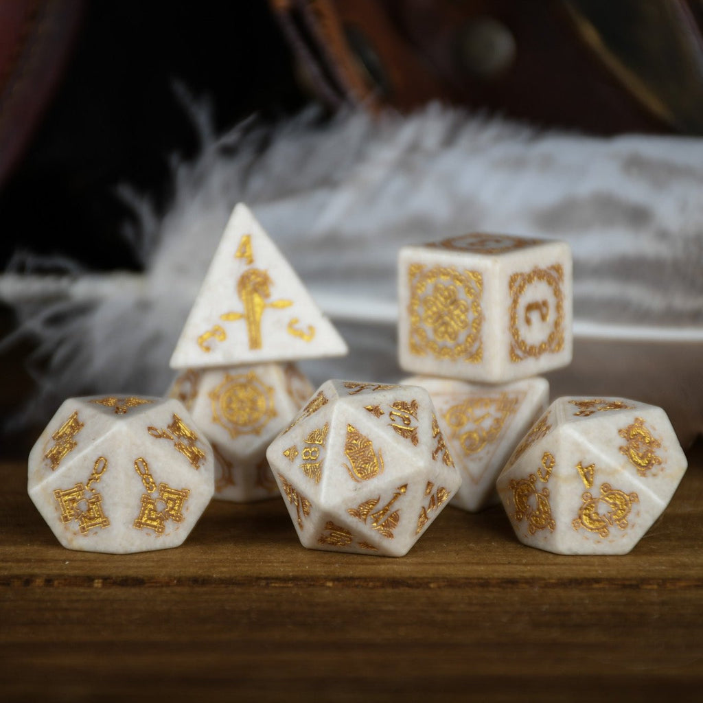 A set of hand-carved white opal stone dice with gold engravings, including various shapes typical for role-playing games like a d4, d6, d8, d10, d12, and d20. The dice are displayed on a wooden surface with a blurred background.