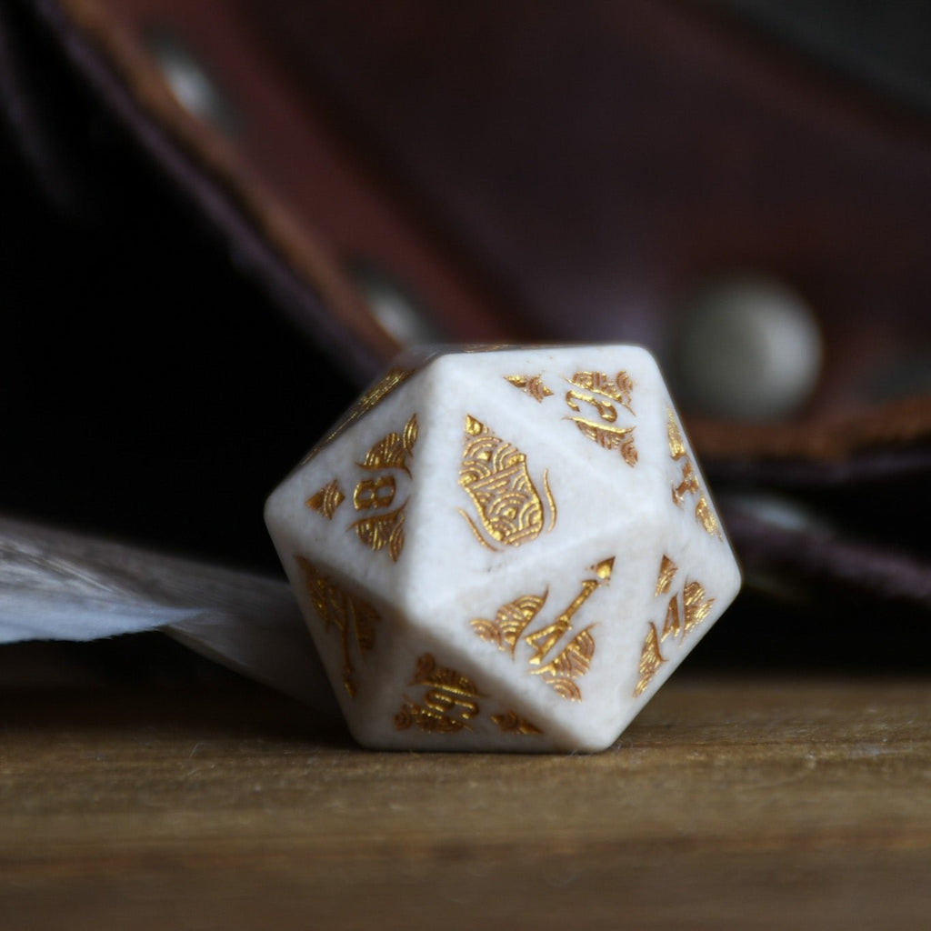 A set of hand-carved white opal stone dice with gold engravings, including various shapes typical for role-playing games like a d4, d6, d8, d10, d12, and d20. The dice are displayed on a wooden surface with a blurred background.