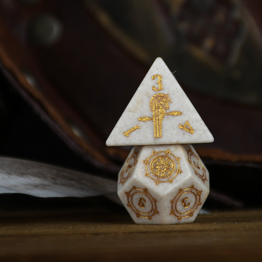 A set of hand-carved white opal stone dice with gold engravings, including various shapes typical for role-playing games like a d4, d6, d8, d10, d12, and d20. The dice are displayed on a wooden surface with a blurred background.