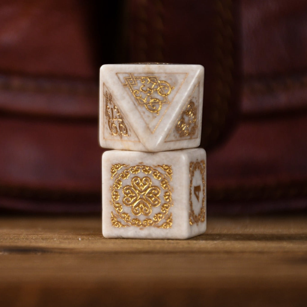 A set of hand-carved white opal stone dice with gold engravings, including various shapes typical for role-playing games like a d4, d6, d8, d10, d12, and d20. The dice are displayed on a wooden surface with a blurred background.