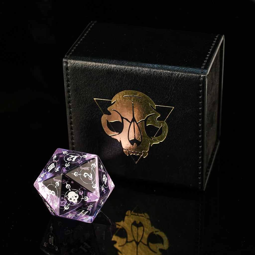Purple glass polyhedral dice with black and white patterns on a black surface with a decorative box.