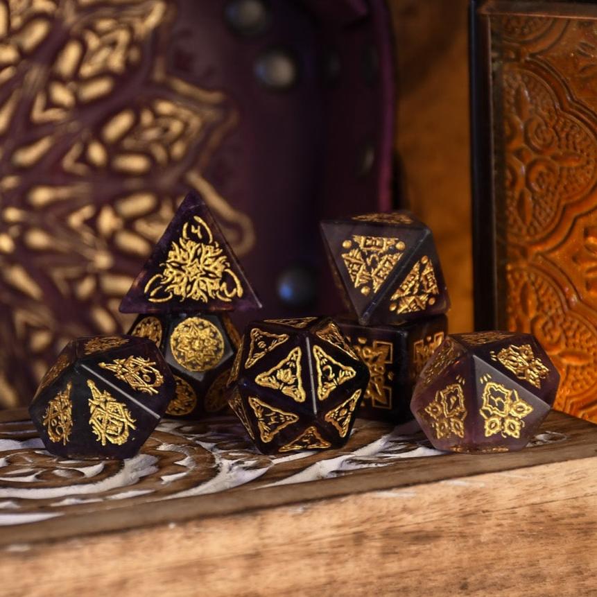 A set of purple fluorite stone dice with gold engravings, displayed on a wooden surface.