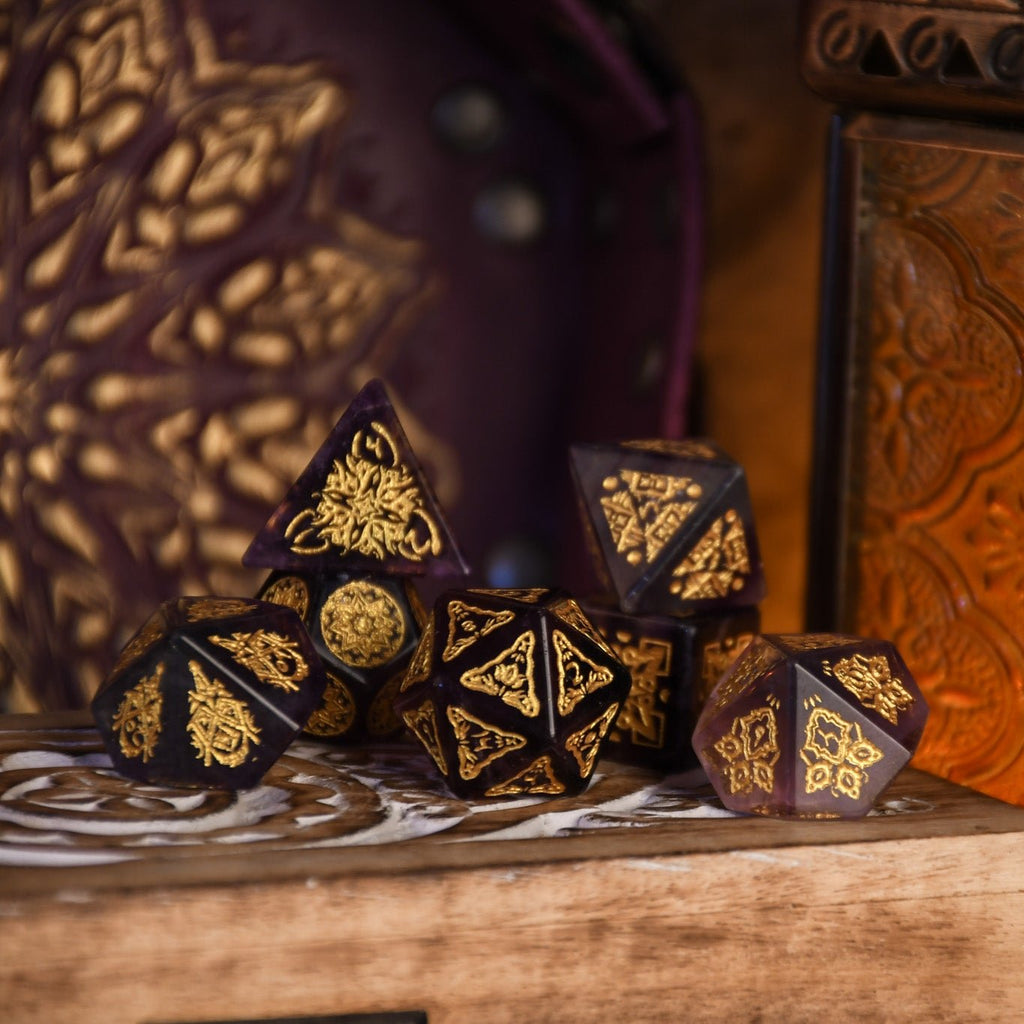 A set of purple fluorite stone dice with gold engravings, displayed on a wooden surface.