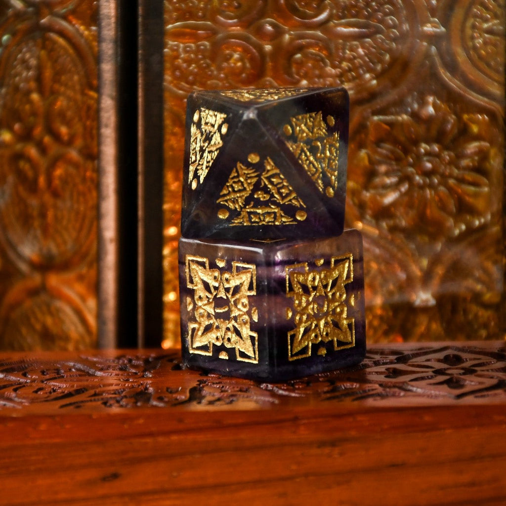 A set of purple fluorite stone dice with gold engravings, displayed on a wooden surface.