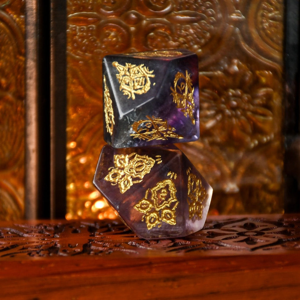 A set of purple fluorite stone dice with gold engravings, displayed on a wooden surface.