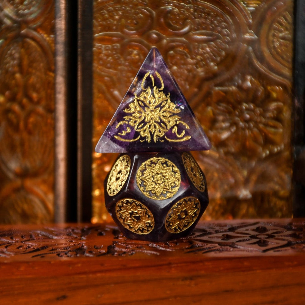 A set of purple fluorite stone dice with gold engravings, displayed on a wooden surface.
