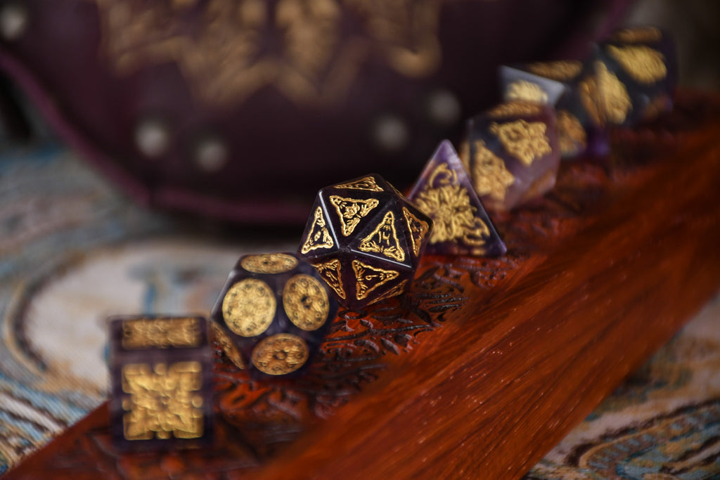A set of purple fluorite stone dice with gold engravings, displayed on a wooden dnd dice vault.