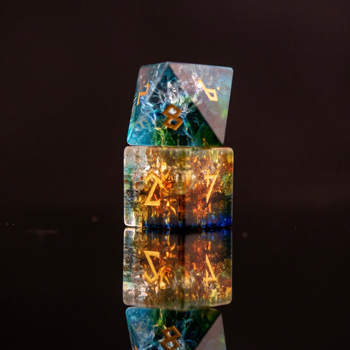 A set of shattered glass dice with engraved numbers, featuring a variety of colors including blue, green, and orange, displayed on a reflective surface.