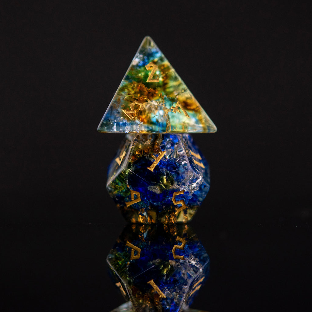 A set of shattered glass dice with engraved numbers, featuring a variety of colors including blue, green, and orange, displayed on a reflective surface.