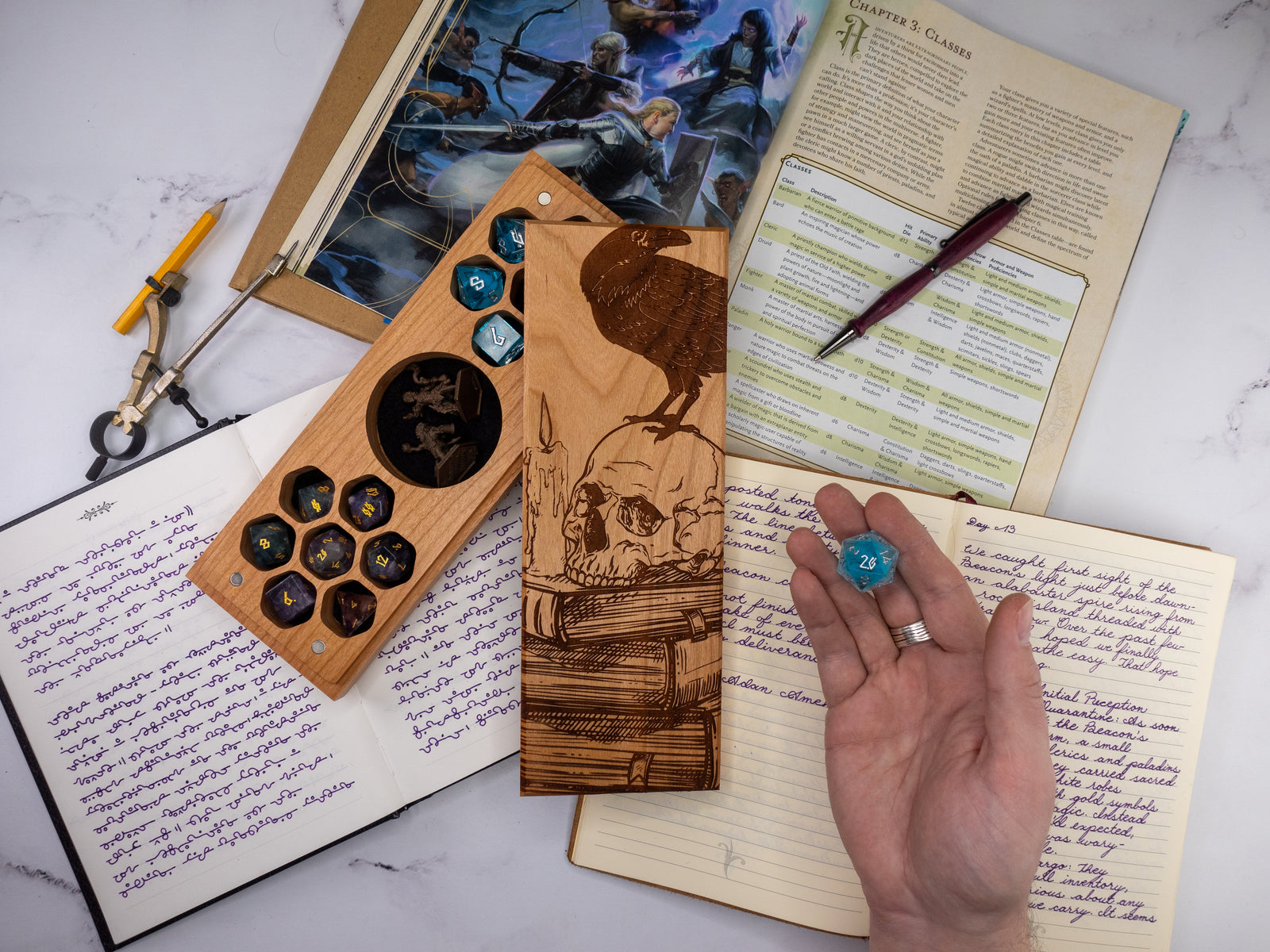 Wooden dice tray with dice, hand holding a blue die, and open notebook on a marble surface