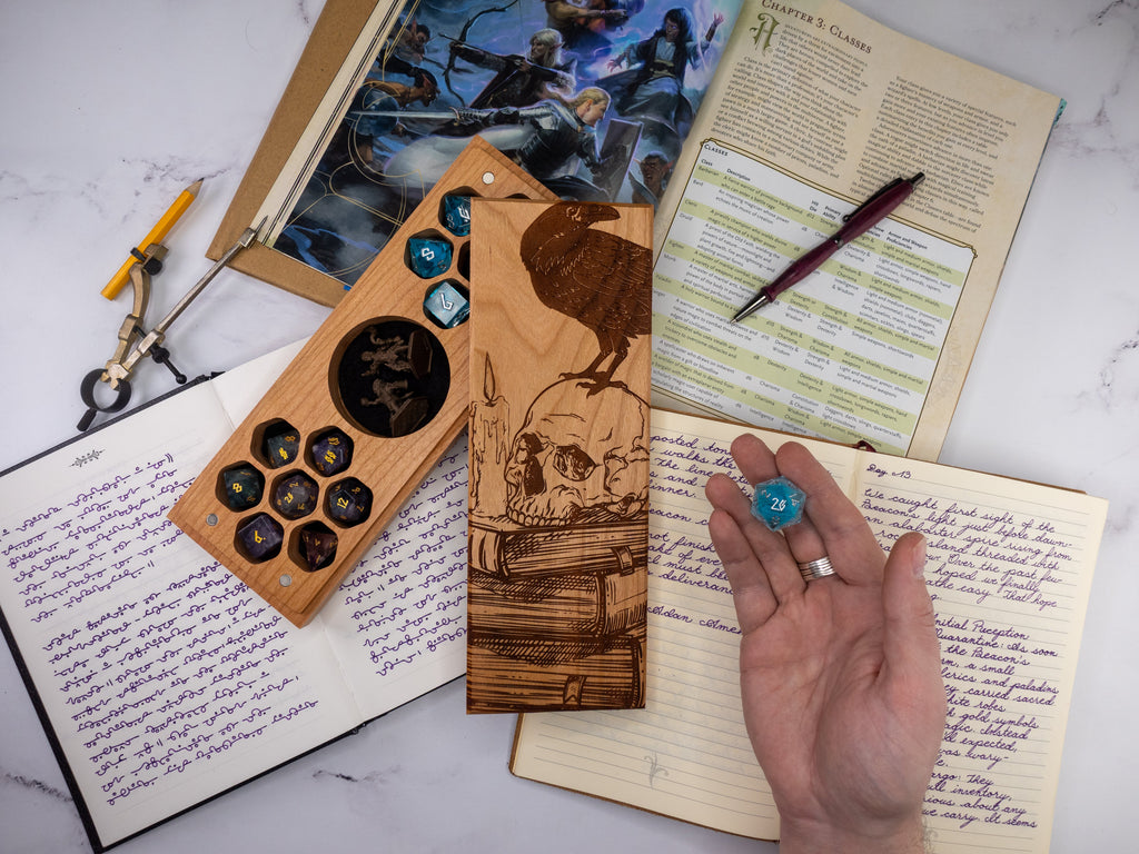 Wooden dice tray with dice, hand holding a blue die, and open notebook on a marble surface