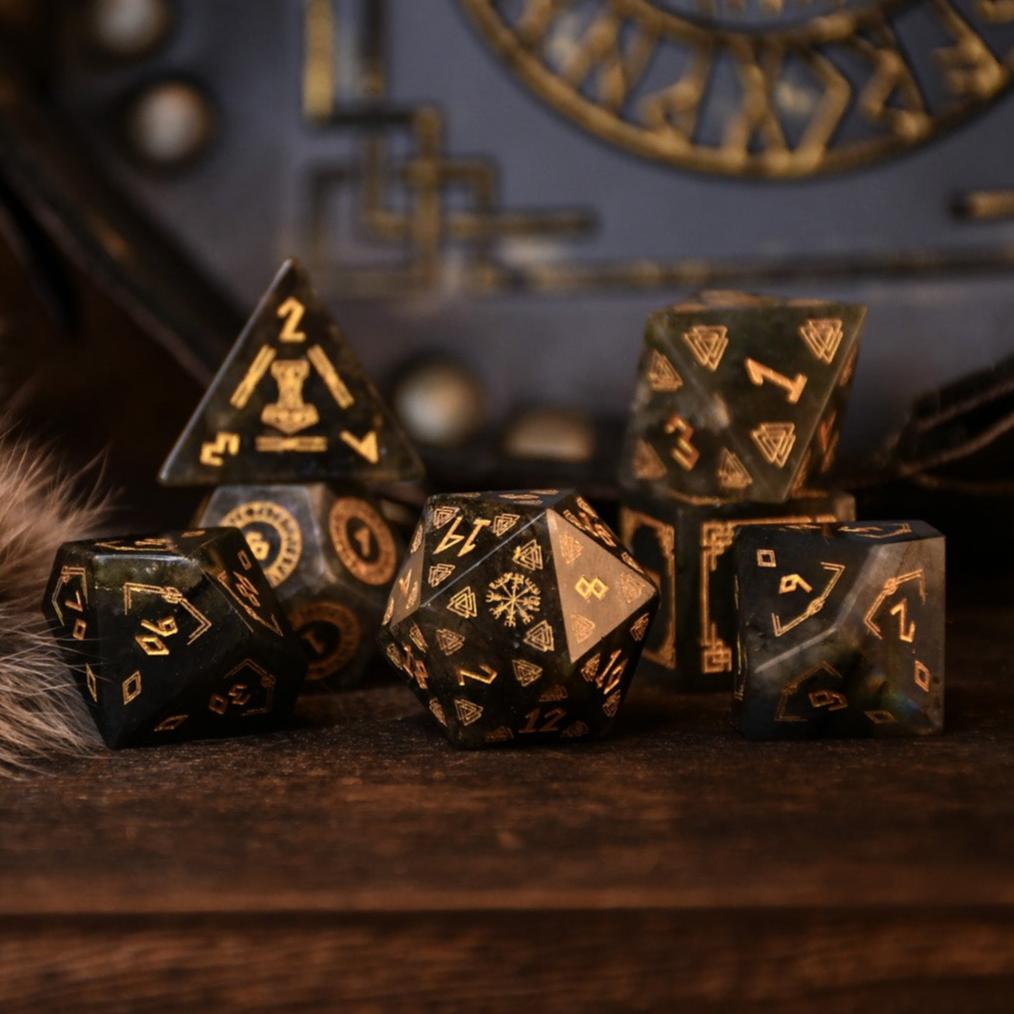 A set of black labradorite stone dice with gold engravings, displayed on a wooden surface. The dice are of various shapes including a four-sided, six-sided, eight-sided, two types of ten-sided, and a twelve-sided die, along with a twenty-sided die.