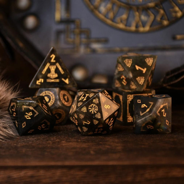 A set of black labradorite stone dice with gold engravings, displayed on a wooden surface. The dice are of various shapes including a four-sided, six-sided, eight-sided, two types of ten-sided, and a twelve-sided die, along with a twenty-sided die.