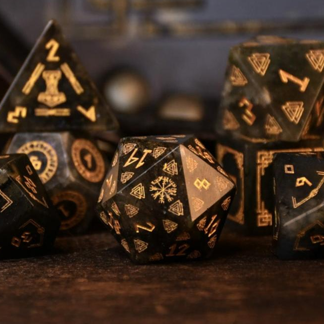 A set of black labradorite stone dice with gold engravings, displayed on a wooden surface. The dice are of various shapes including a four-sided, six-sided, eight-sided, two types of ten-sided, and a twelve-sided die, along with a twenty-sided die.