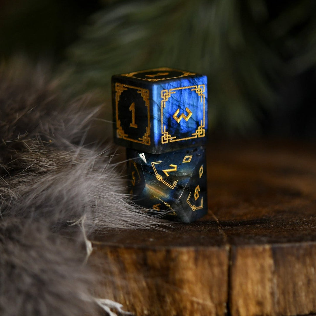A set of black labradorite stone dice with gold engravings, displayed on a wooden surface. The dice are of various shapes including a four-sided, six-sided, eight-sided, two types of ten-sided, and a twelve-sided die, along with a twenty-sided die.