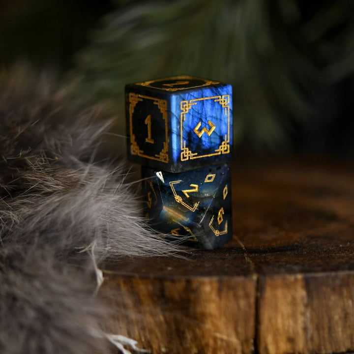 A set of black labradorite stone dice with gold engravings, displayed on a wooden surface. The dice are of various shapes including a four-sided, six-sided, eight-sided, two types of ten-sided, and a twelve-sided die, along with a twenty-sided die.