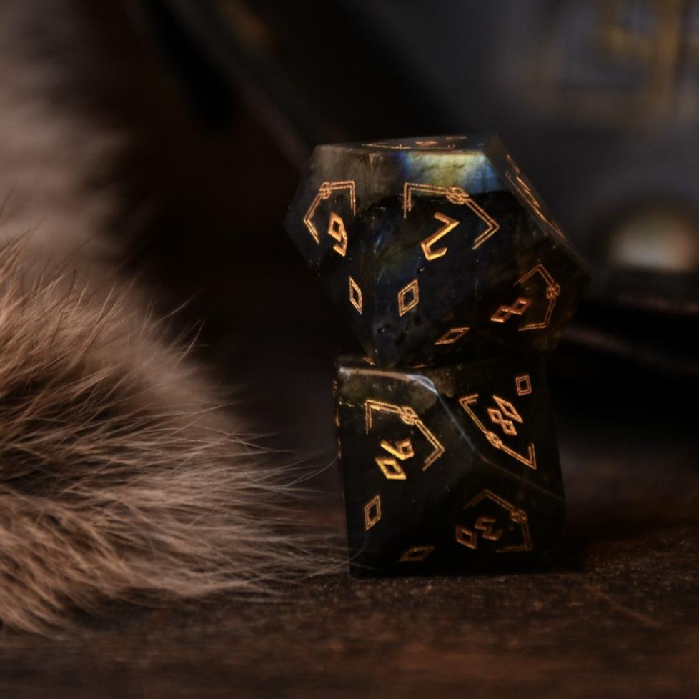 A set of black labradorite stone dice with gold engravings, displayed on a wooden surface. The dice are of various shapes including a four-sided, six-sided, eight-sided, two types of ten-sided, and a twelve-sided die, along with a twenty-sided die.