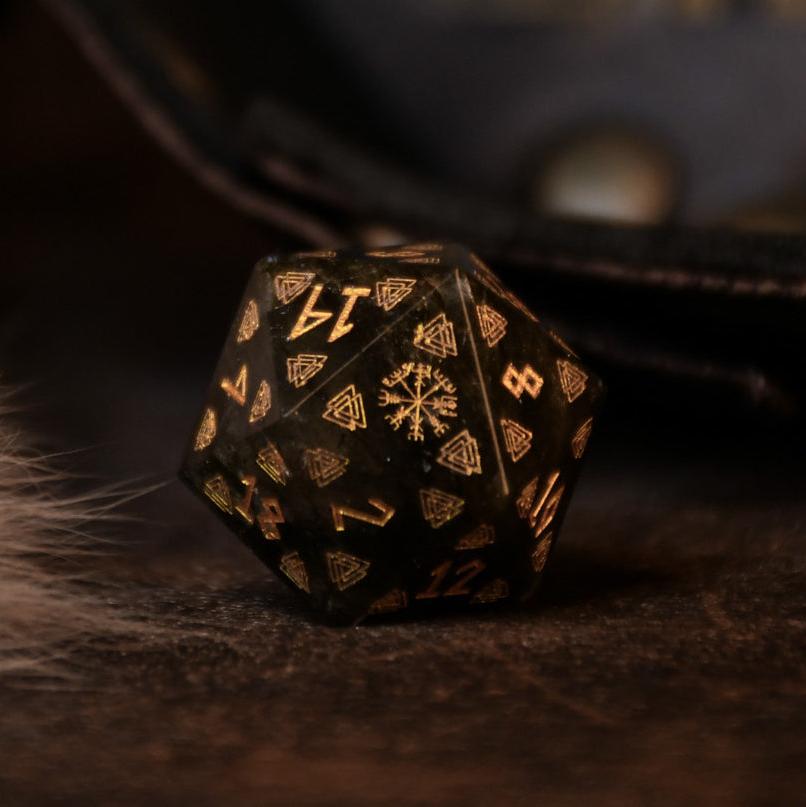 A set of black labradorite stone dice with gold engravings, displayed on a wooden surface. The dice are of various shapes including a four-sided, six-sided, eight-sided, two types of ten-sided, and a twelve-sided die, along with a twenty-sided die.