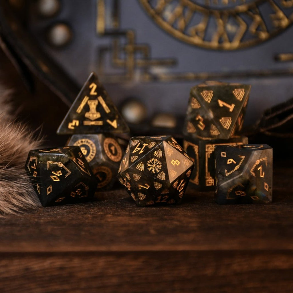 A set of black labradorite stone dice with gold engravings, displayed on a wooden surface. The dice are of various shapes including a four-sided, six-sided, eight-sided, two types of ten-sided, and a twelve-sided die, along with a twenty-sided die.