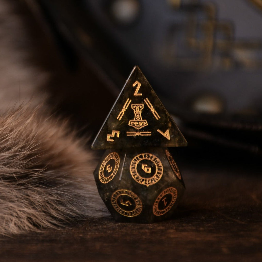 A set of black labradorite stone dice with gold engravings, displayed on a wooden surface. The dice are of various shapes including a four-sided, six-sided, eight-sided, two types of ten-sided, and a twelve-sided die, along with a twenty-sided die.