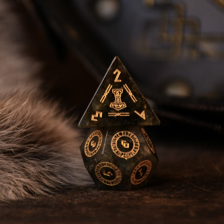 A set of black labradorite stone dice with gold engravings, displayed on a wooden surface. The dice are of various shapes including a four-sided, six-sided, eight-sided, two types of ten-sided, and a twelve-sided die, along with a twenty-sided die.