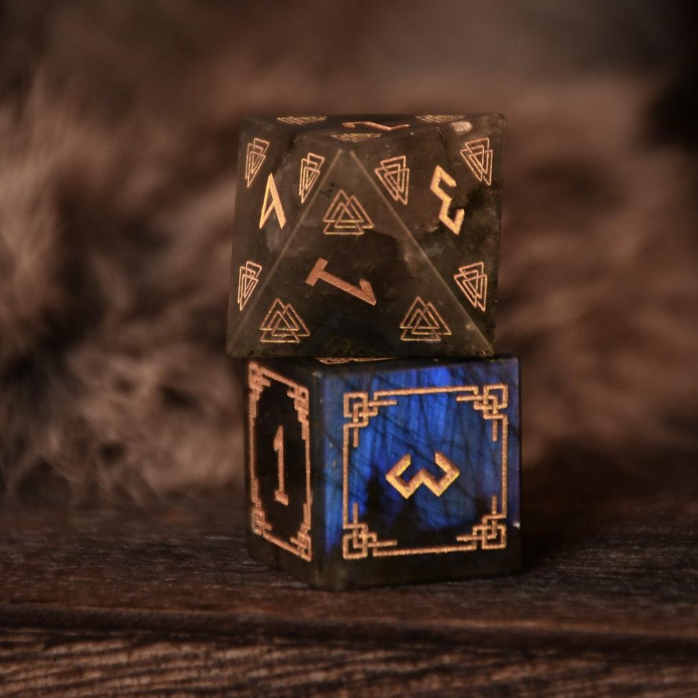 A set of black labradorite stone dice with gold engravings, displayed on a wooden surface. The dice are of various shapes including a four-sided, six-sided, eight-sided, two types of ten-sided, and a twelve-sided die, along with a twenty-sided die.