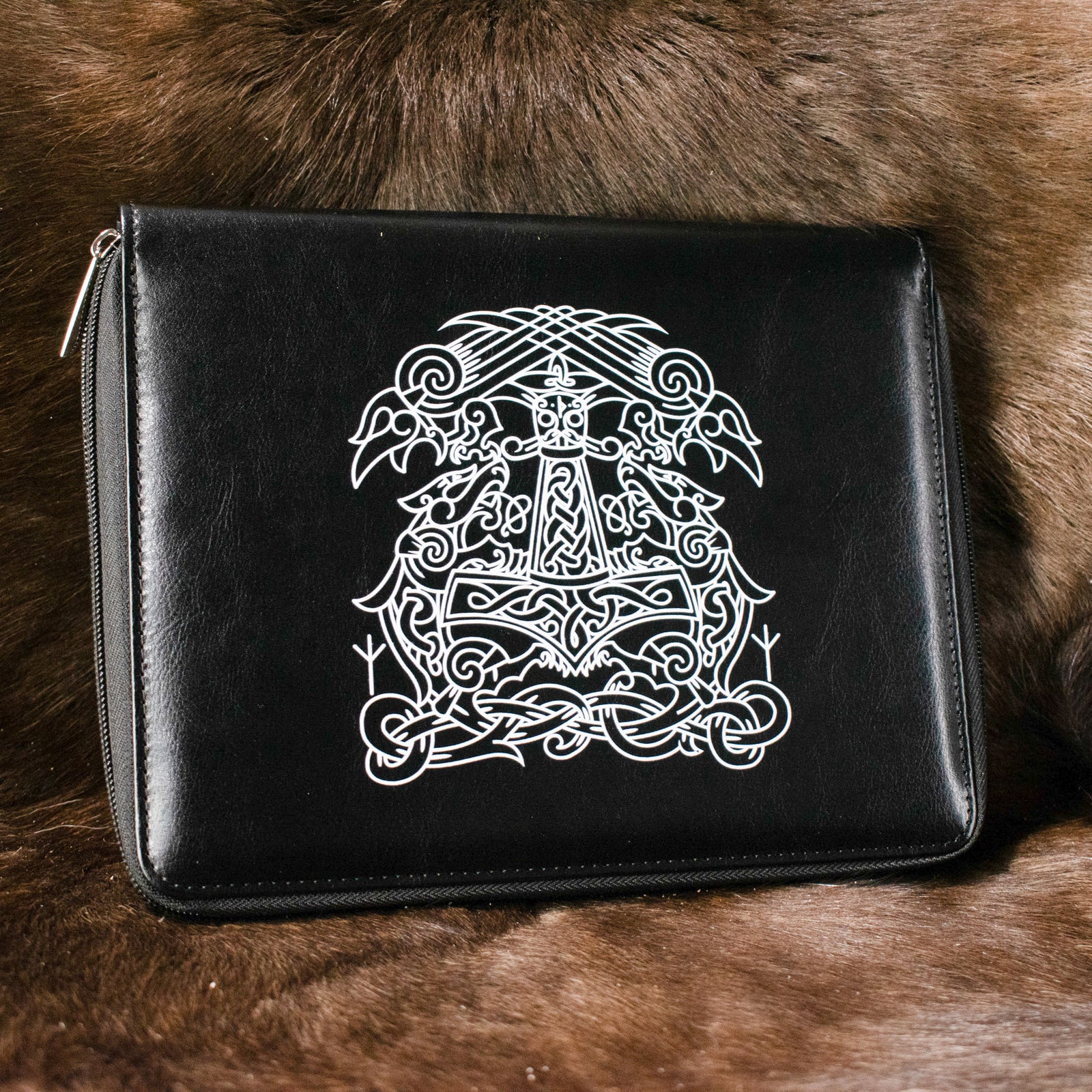 A black poly-leather zippered dice case with a white printed Norse runic symbol on the front, placed on a furry surface.
