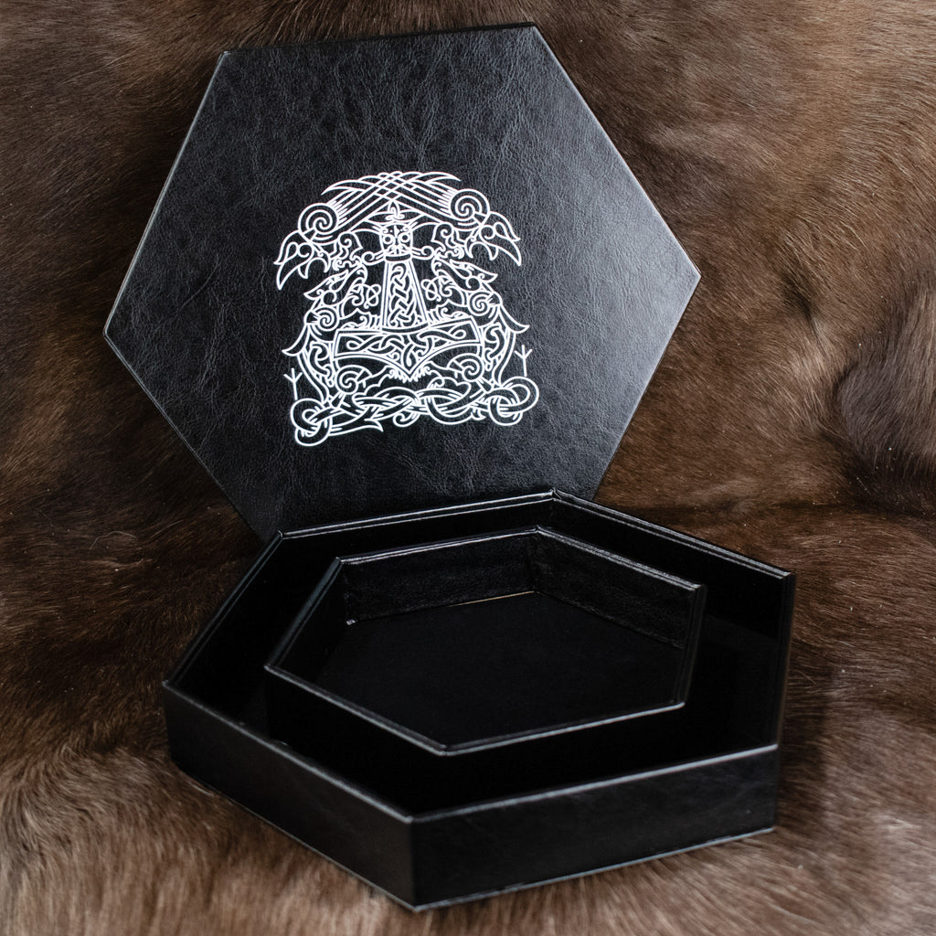 A black hexagonal dice tray with a white Norse-inspired pattern on the lid, placed on a furry surface.
