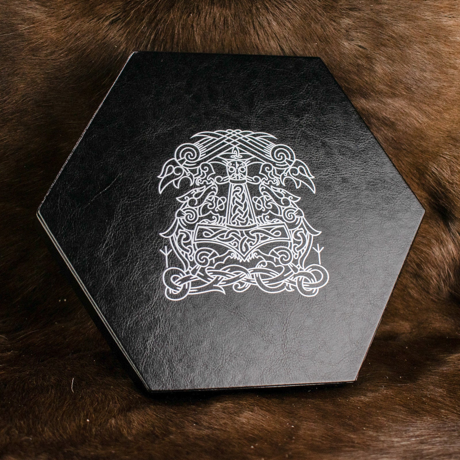 A black hexagonal dice tray with a white Norse-inspired pattern on the lid, placed on a furry surface.