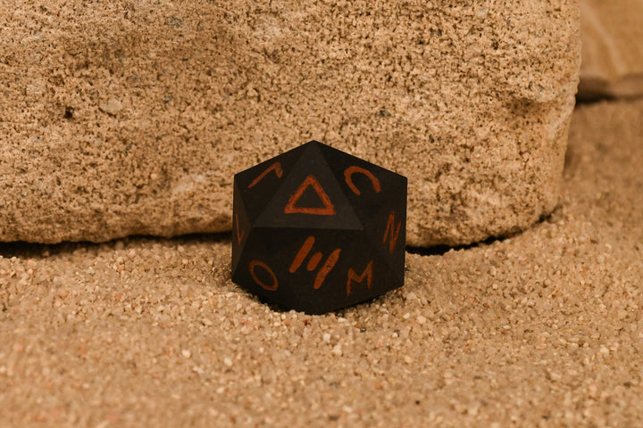 A 30mm black serpentine stone d20 dice with ancient Greek letters inscribed on each face, positioned on a textured surface.