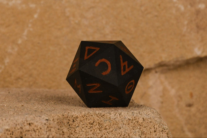 A 30mm black serpentine stone d20 dice with ancient Greek letters inscribed on each face, positioned on a textured surface.
