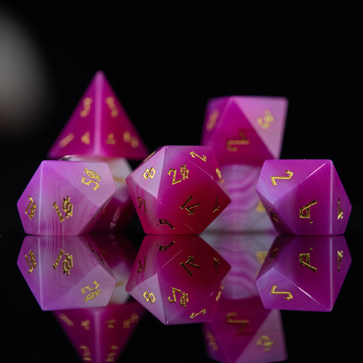 A set of pink agate stone dice with engraved numbers, polished and displayed on a reflective surface.