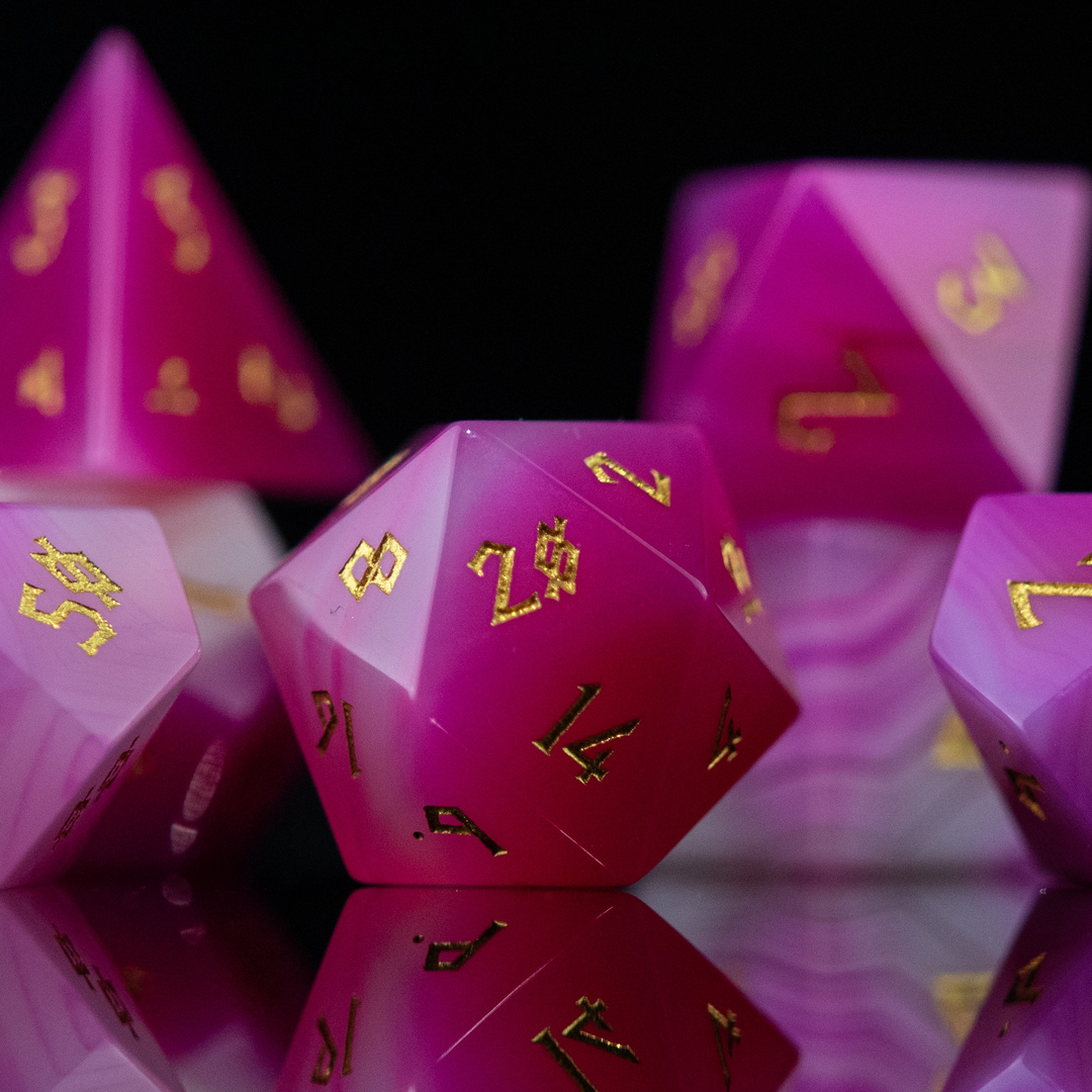 A set of pink agate stone dice with engraved numbers, polished and displayed on a reflective surface.