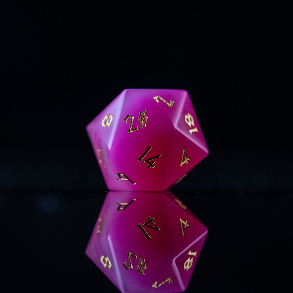 A set of pink agate stone dice with engraved numbers, polished and displayed on a reflective surface.