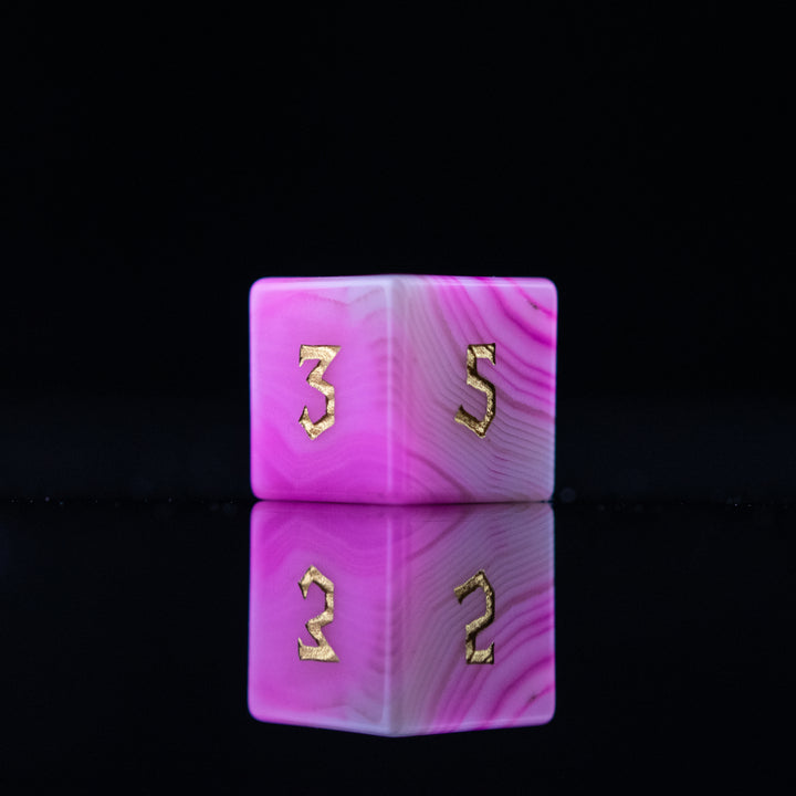 A set of pink agate stone dice with engraved numbers, polished and displayed on a reflective surface.