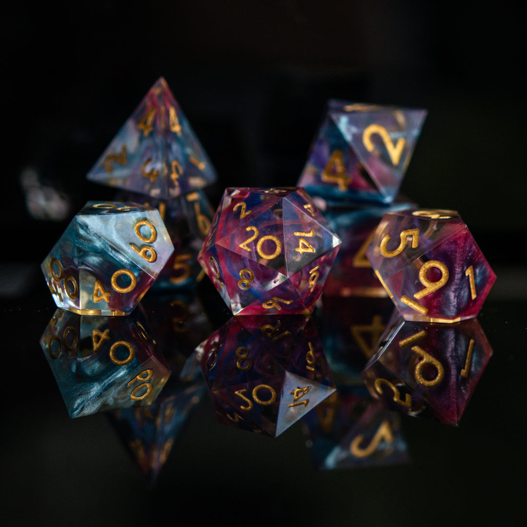 A set of colorful sharp-edged resin dice with hand-painted numbers, featuring a multicolor finish with gold numbers, displayed on a reflective surface.