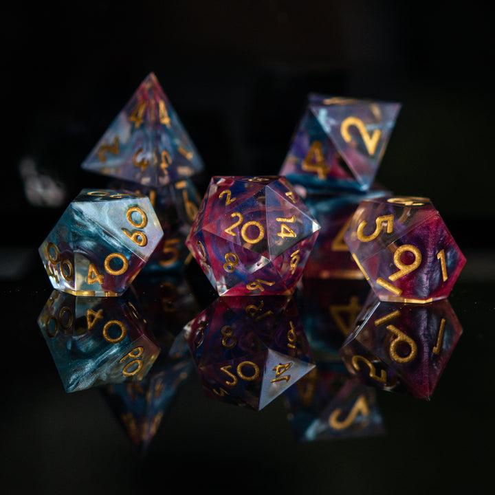 A set of colorful sharp-edged resin dice with hand-painted numbers, featuring a multicolor finish with gold numbers, displayed on a reflective surface.