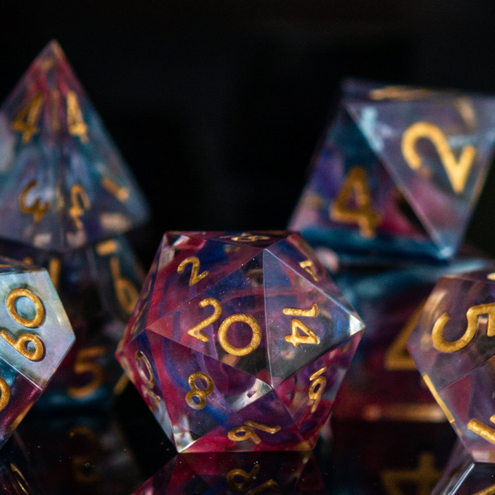 A set of colorful sharp-edged resin dice with hand-painted numbers, featuring a multicolor finish with gold numbers, displayed on a reflective surface.