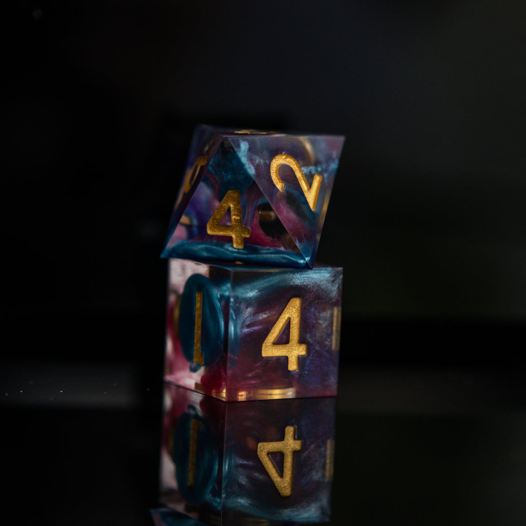 A set of colorful sharp-edged resin dice with hand-painted numbers, featuring a multicolor finish with gold numbers, displayed on a reflective surface.
