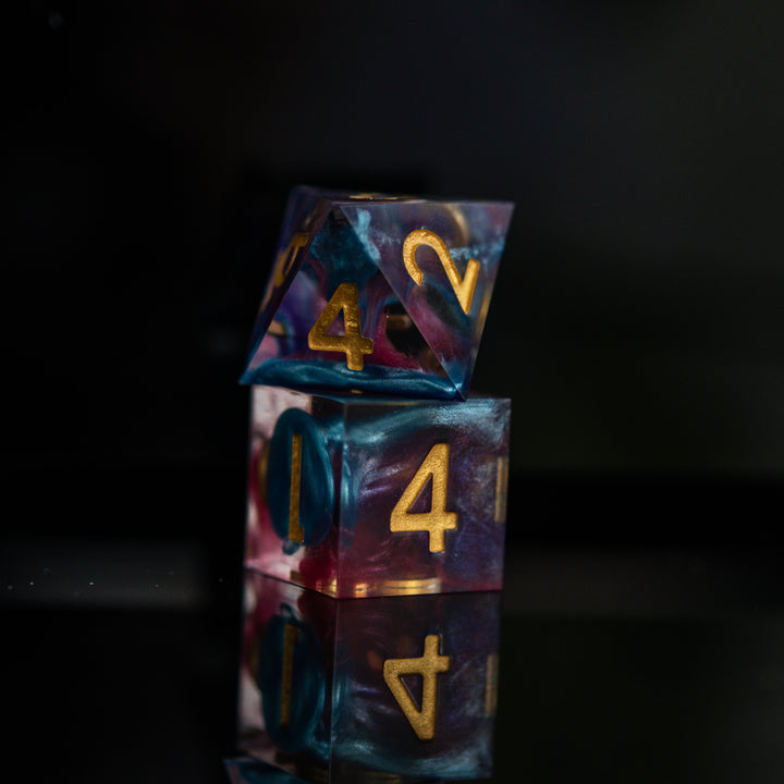 A set of colorful sharp-edged resin dice with hand-painted numbers, featuring a multicolor finish with gold numbers, displayed on a reflective surface.