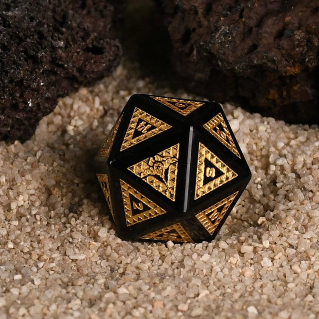 A set of black obsidian stone dice with gold engravings, displayed on a sandy surface with a green plant in the background.