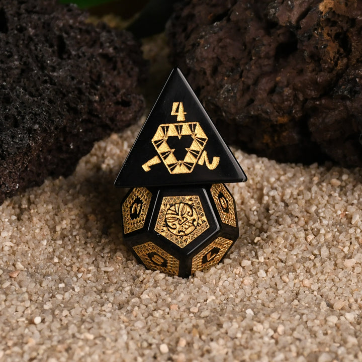 A set of black obsidian stone dice with gold engravings, displayed on a sandy surface with a green plant in the background.