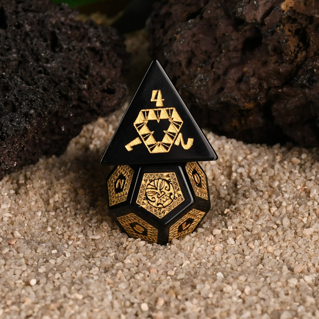 A set of black obsidian stone dice with gold engravings, displayed on a sandy surface with a green plant in the background.