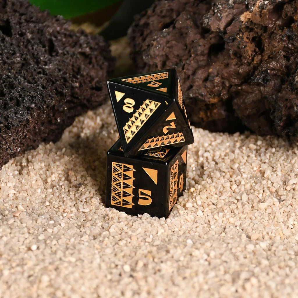 A set of black obsidian stone dice with gold engravings, displayed on a sandy surface with a green plant in the background.