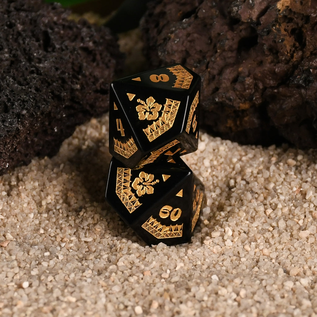 A set of black obsidian stone dice with gold engravings, displayed on a sandy surface with a green plant in the background.