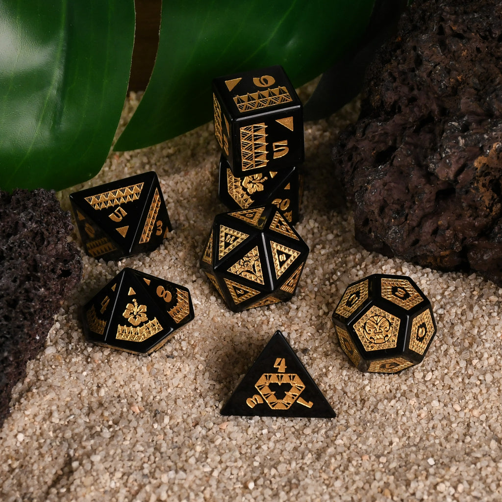 A set of black obsidian stone dice with gold engravings, displayed on a sandy surface with a green plant in the background.